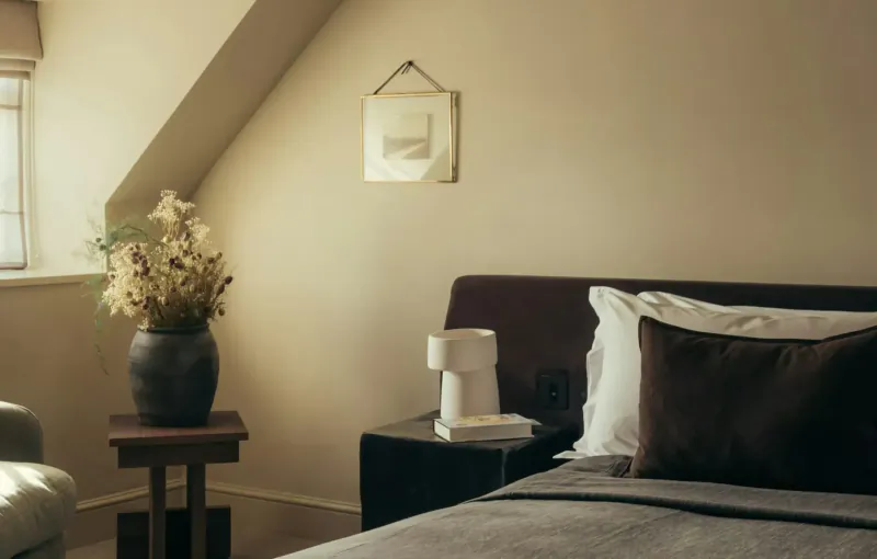 Cozy attic bedroom with sloped ceiling, dried flowers on side table, lamp, books on nightstand, and bed with pillows.