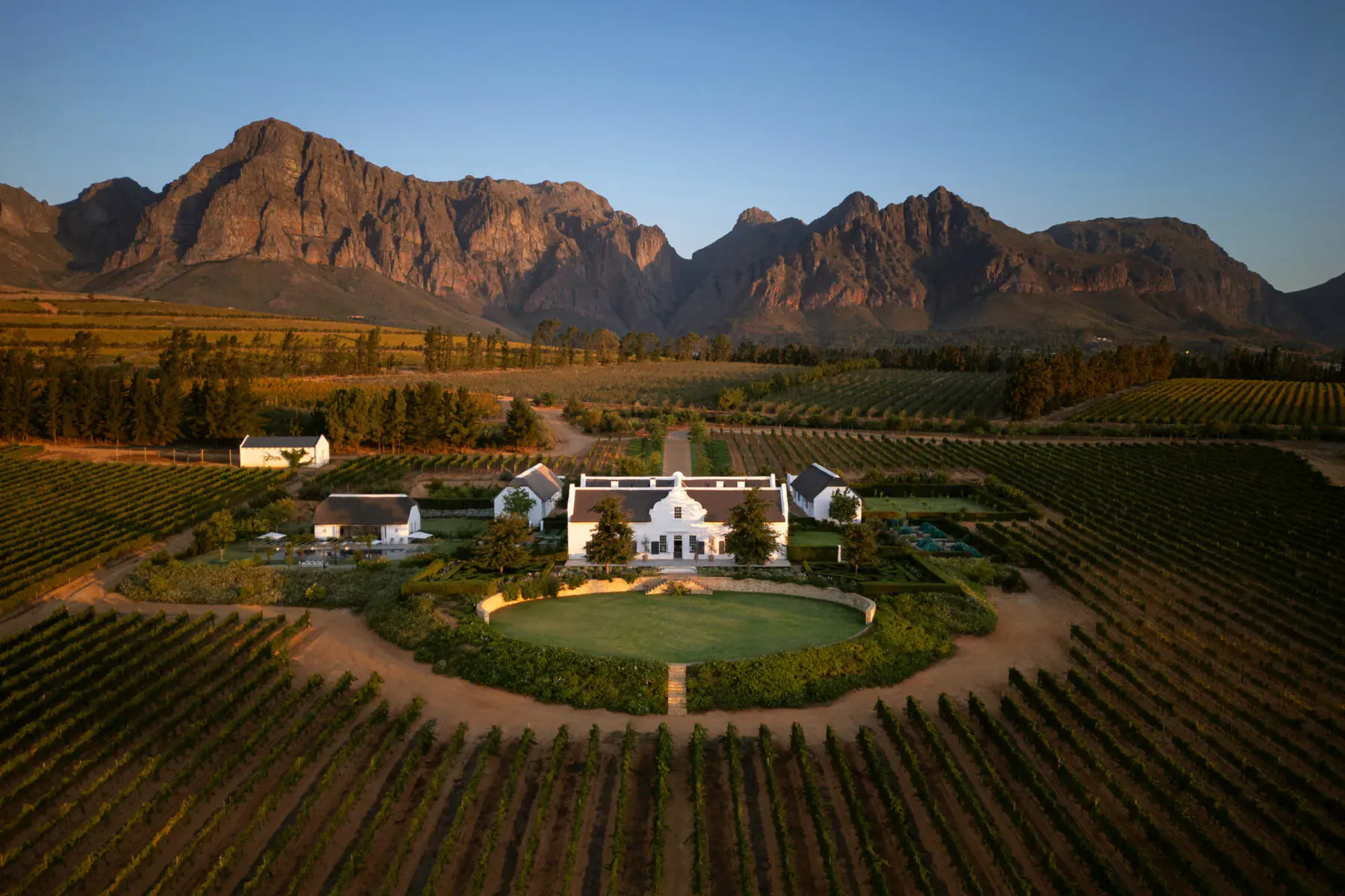 Aerial view of Brookdale Wine Estate in Paarl: white Cape Dutch manor amid vineyards, gardens, and rugged mountains at sunset.