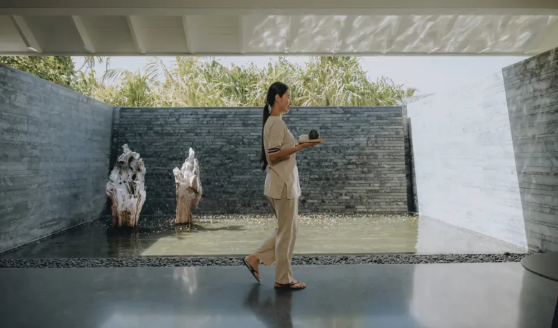 Woman in beige uniform holding phone walks past stone sculptures in modern Maldives resort courtyard