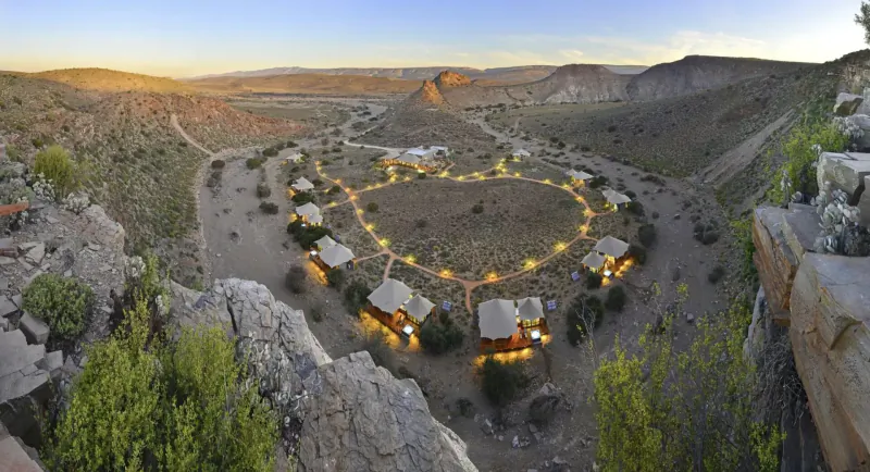 Panoramic sunset view of circular glamping tents lit up in a rugged canyon valley, ethical savanna travel.
