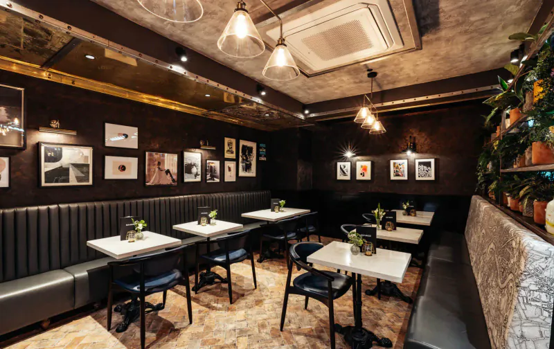 Elegant Six by Nico Canary Wharf gallery dining room with Ancient Rome theme, plush black benches, white tables with greenery, warm pendant lights, art on textured walls.