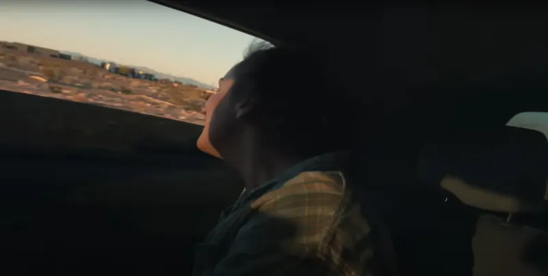 Black driver in plaid shirt looks up from IONIQ 5 robotaxi, cityscape at sunset visible through window