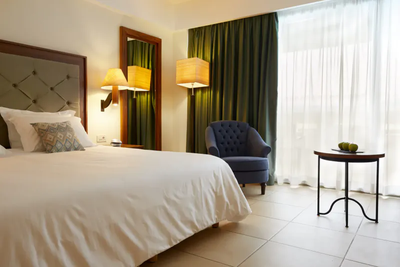 Luxurious hotel bedroom with white bed, green curtains, lamps, blue armchair, side table with fruit bowl, and balcony view.