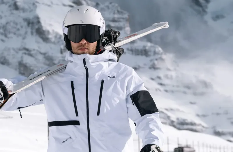Man in white Oakley ski jacket and helmet holding skis on snowy mountain slope
