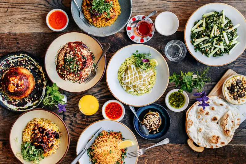 Overhead view of Bahraini dishes on wooden table: curries, rice, naan, salads, dips, and sauces.