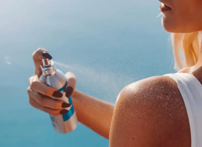 Woman with blonde hair spraying SunKiss suncare from silver aerosol bottle on her shoulder outdoors