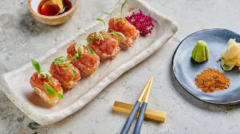 Five tuna nigiri sushi pieces topped with microgreens on white rectangular plate, with chopsticks, ginger, wasabi, and garnishes