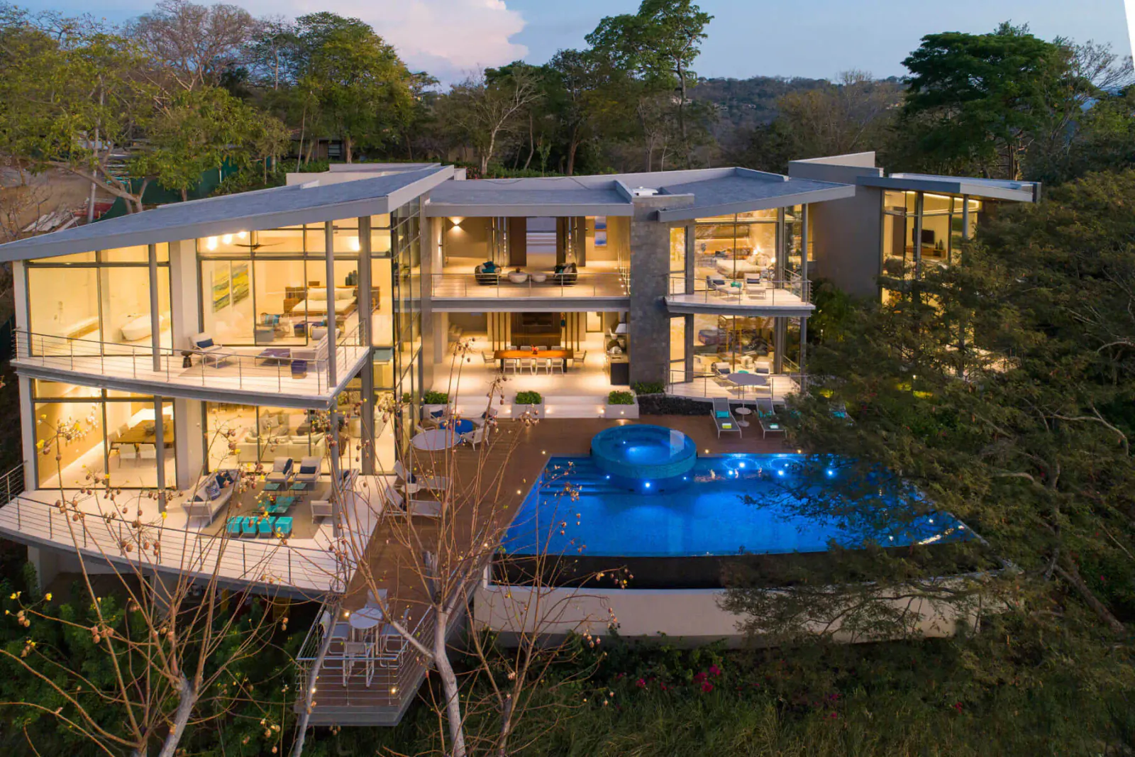 Aerial view of modern luxury multi-level home with glass walls, infinity pool, and tropical surroundings at dusk