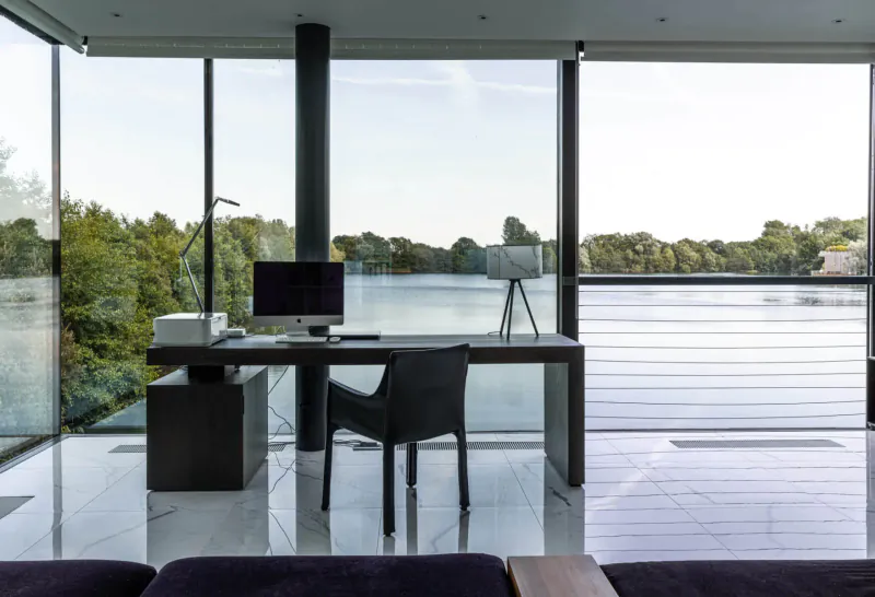 Modern black desk with computer and lamp in glasshouse office overlooking serene lake and trees