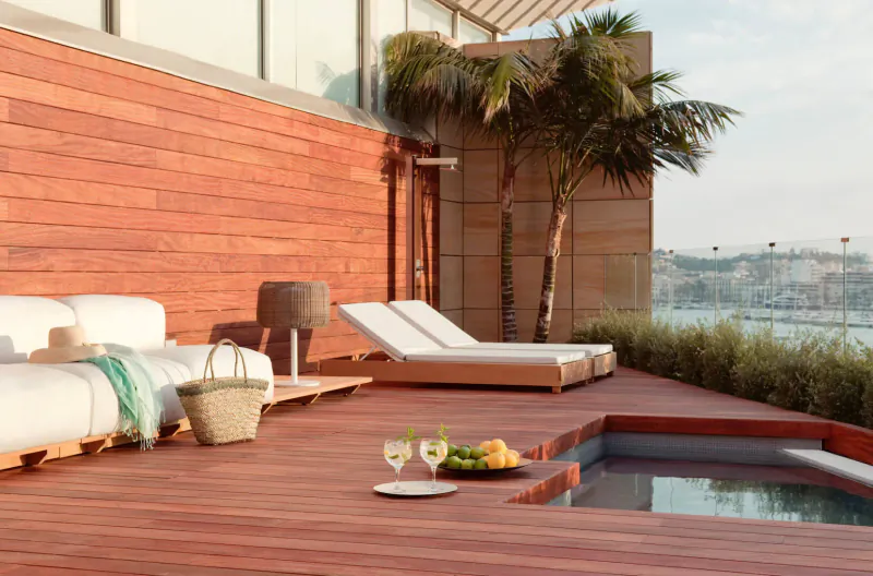 Luxury rooftop deck with white loungers, wine glasses, fruit plate by infinity pool, palms, red wood, sea view.