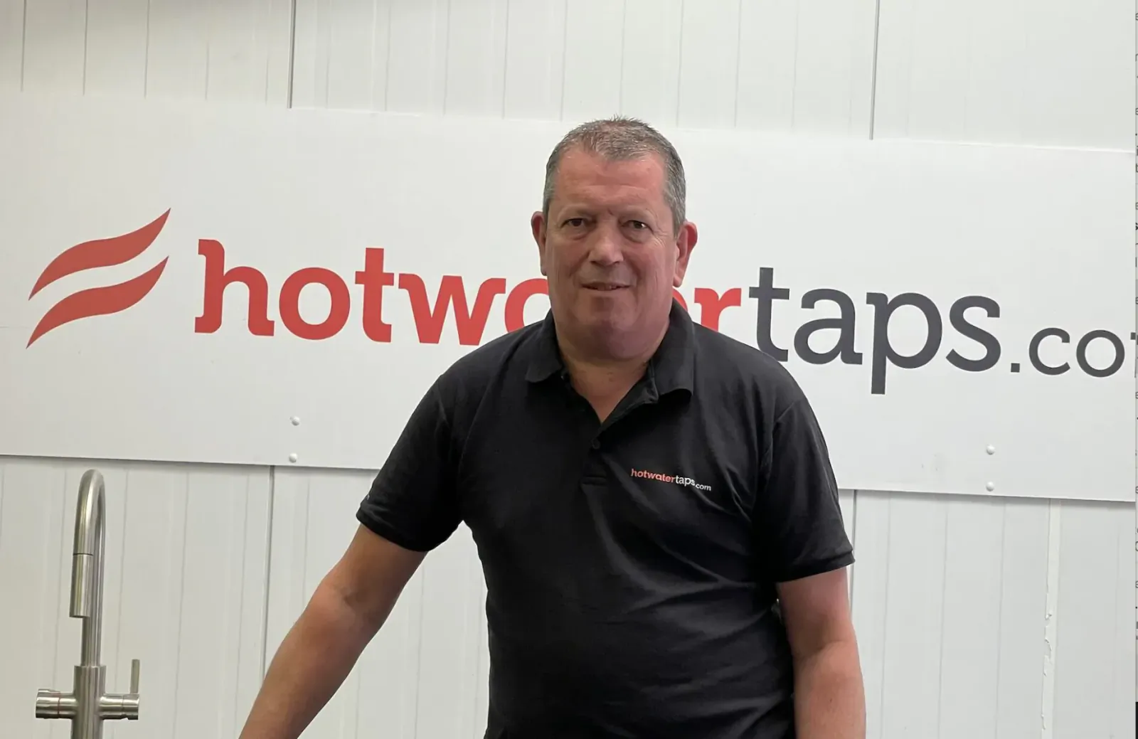 Darren Cooper, GM of Hot Water Taps (hotwatertaps.com), smiling in black polo before company logo wall.