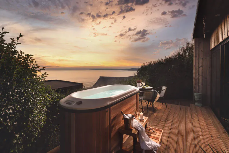 Wooden hot tub on deck of clifftop Cornwall cabin at sunset, overlooking ocean with shrubs and chairs.