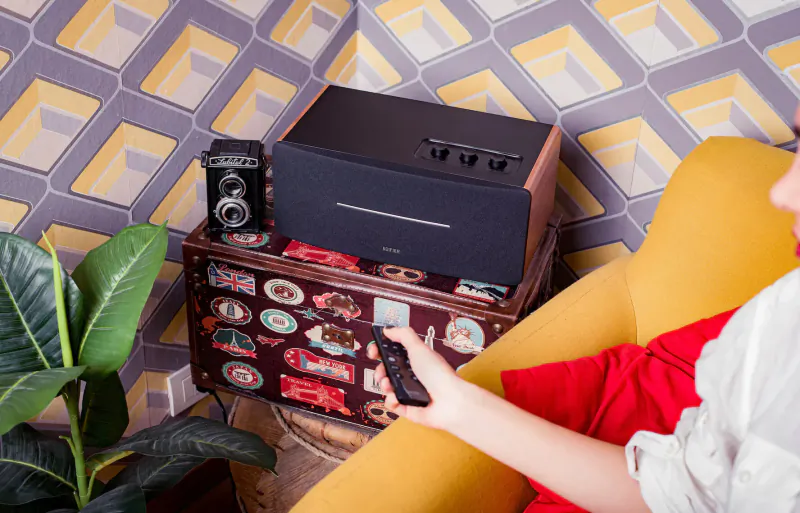 Woman holding remote for new Edifier D12 black wood Bluetooth speaker on vintage suitcase, yellow chair, plants, geometric wall.