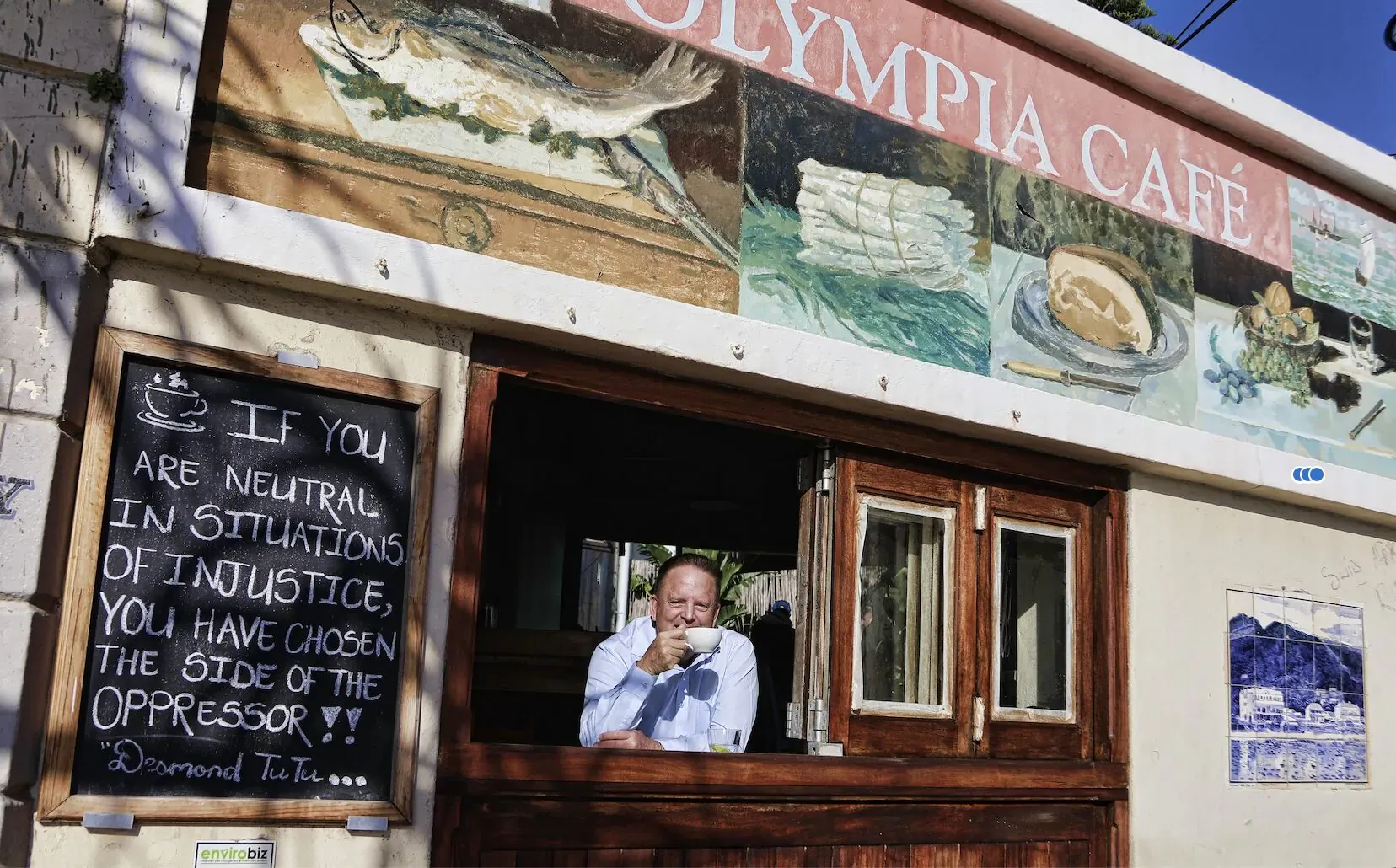 Man in white shirt sips coffee at Olympia's Cafe window in Kalk Bay; mural of fish and plates; chalkboard: 'I live neutral in justice... on the oppressed side'