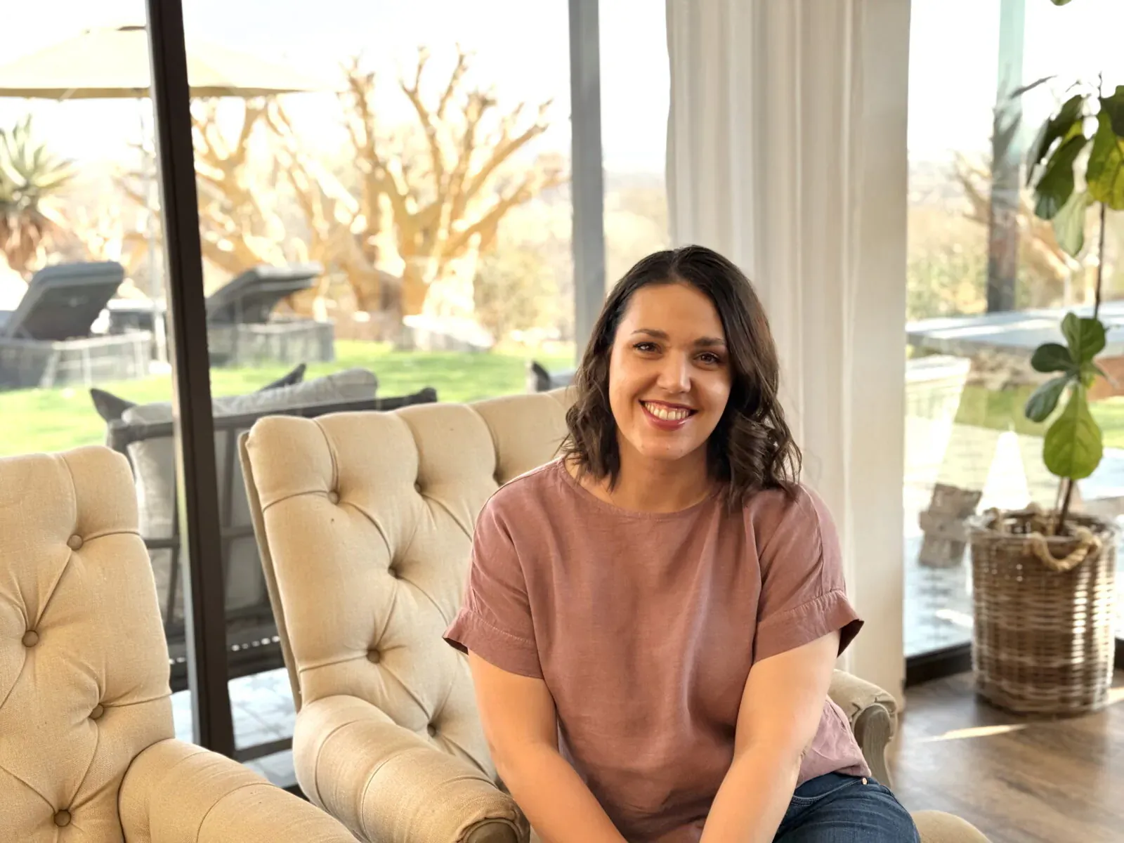 Marquerite Muller, manager of The Otjiwa Collection, smiling on cream armchair in sunlit room with garden view