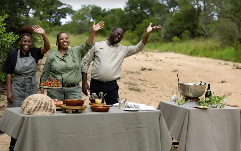 Three smiling staff waving arms beside outdoor bush table with salads, fruits, and drinks at game lodge