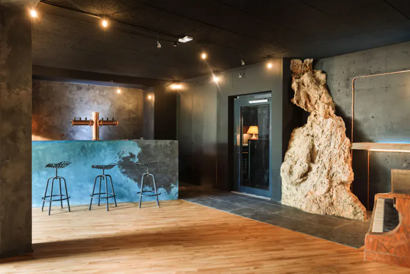 Modern bar area in Lou Calen gallery with teal counter, blue stools, large rock sculpture, cross wall art, warm lighting