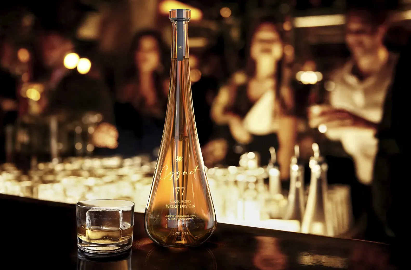 Tall amber Grey Goose bottle and whiskey glass on a dimly lit bar with blurred patrons in background