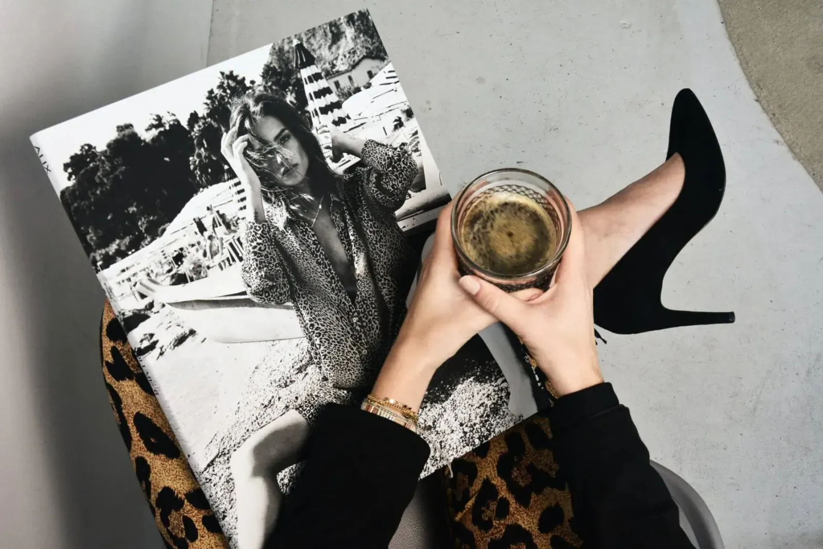 Woman in black holding coffee glass and leopard chair arm, reading open black-and-white fashion photo book with model.