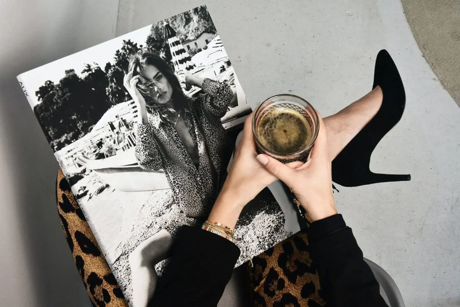 Woman in black holding coffee glass and leopard chair arm, reading open black-and-white fashion photo book with model.
