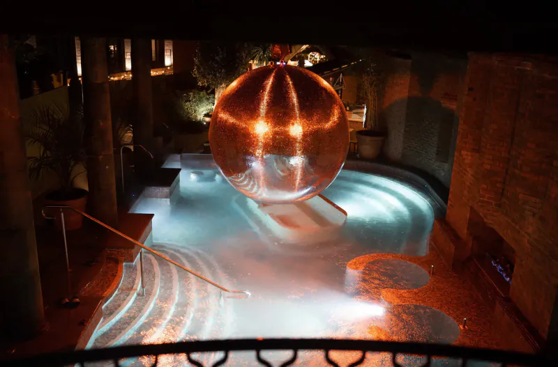 Glowing orange mirrored sphere floating in illuminated indoor pool at night, surrounded by stone architecture and plants