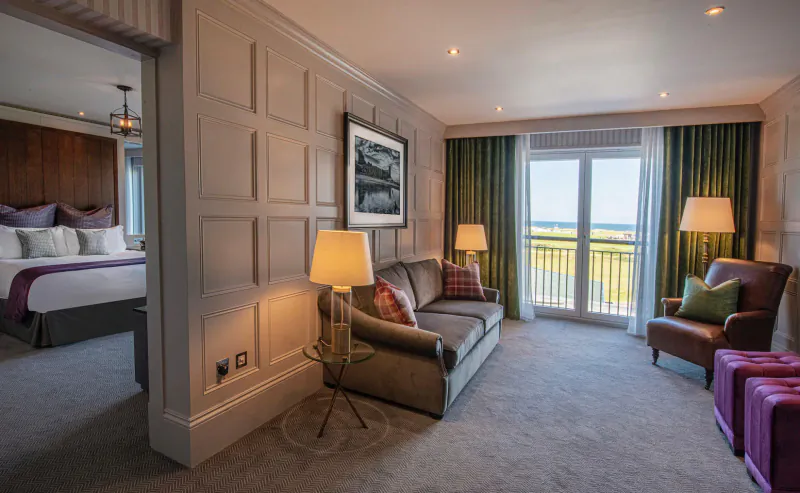Elegant hotel suite with king bed, plush sofa, armchairs, sea view balcony, and golf course print.