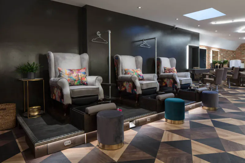 Modern pedicure station in Young LDN Medispa with gray armchairs, colorful cushions, ottomans, and black wall with coat rack.