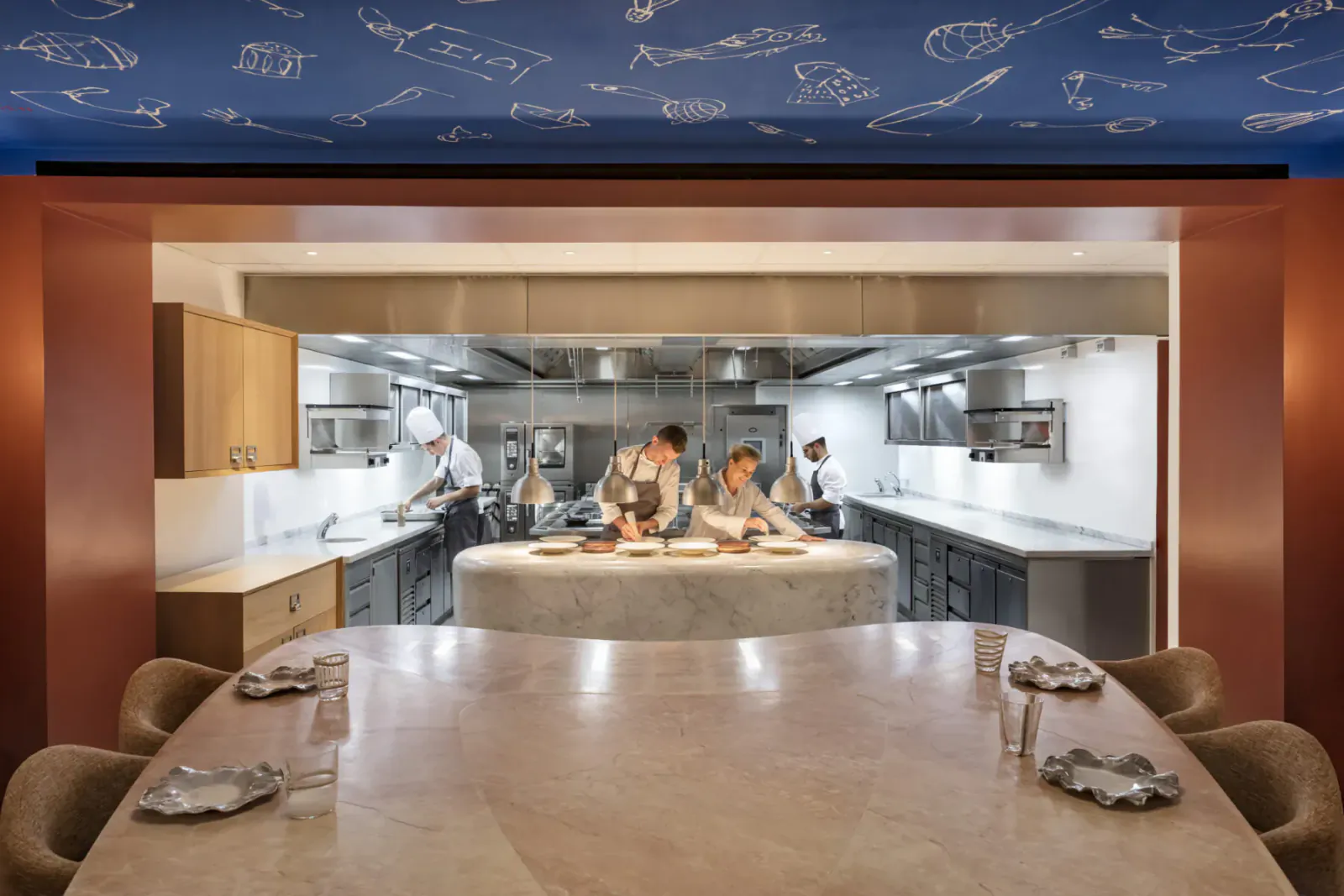 Modern open kitchen with chefs preparing dishes behind a large marble dining table, starry ceiling above.