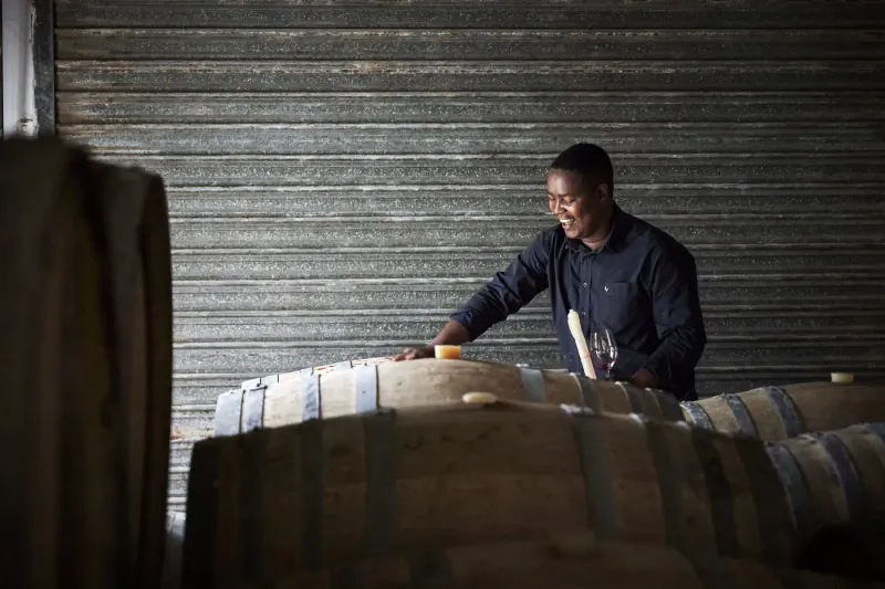 Smiling Black man in dark shirt inspecting wooden wine barrels in dimly lit warehouse