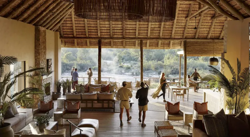 Spacious thatched savanna lodge interior with lounge seating, guests walking, and river view through large windows.