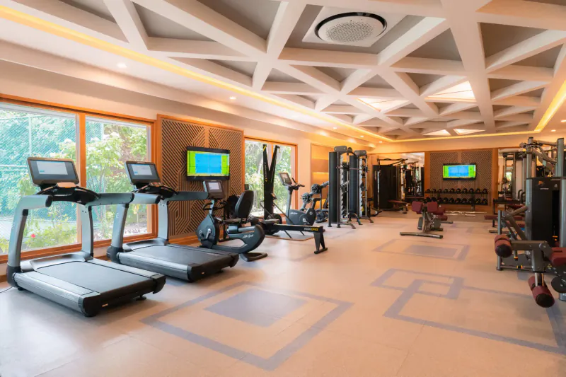Modern gym at Hideaway Beach Resort in Maldives with treadmills, weights, ellipticals, large windows, and TV screens.
