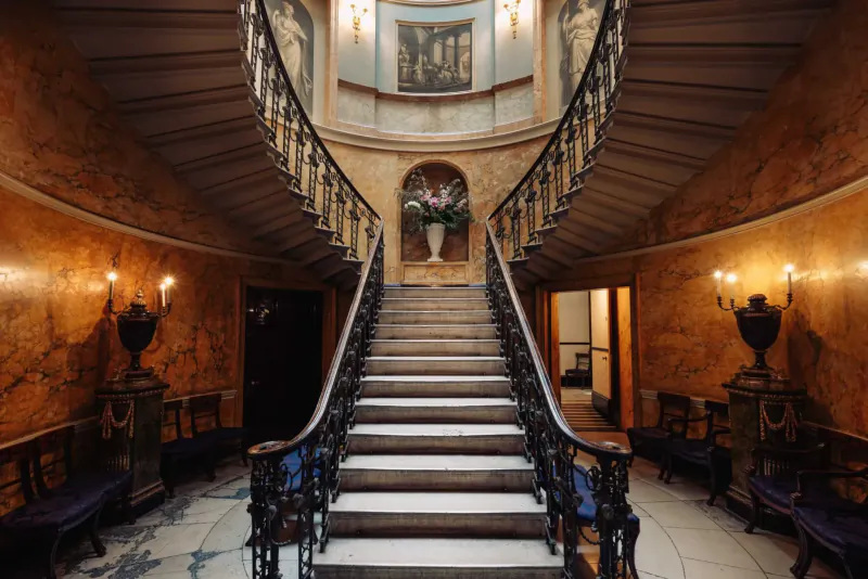 Grand curved wrought-iron staircase in opulent Home House club, with vase of flowers, gilded walls, and wall lamps.