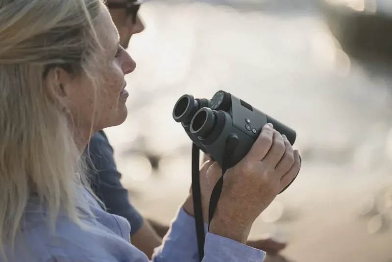 Woman with blonde hair holds Swarovski smart binoculars, peering through them on beach with man beside her