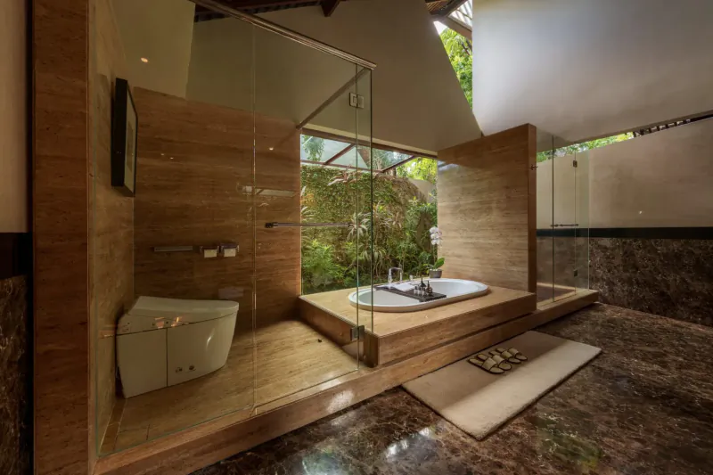 Luxurious modern bathroom in Amétis Villa Bali with wooden walls, glass shower, elevated soaking tub, lush garden view