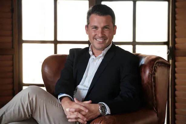 Julián Romaguera smiling confidently in navy suit and white shirt, seated in leather armchair by window with wooden shutters