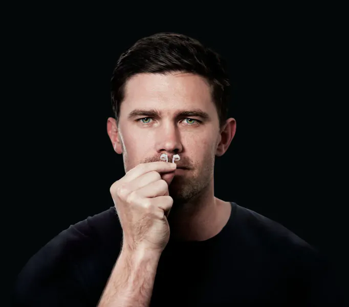 Man with short dark hair holding small white nose clip to his nostril, black background