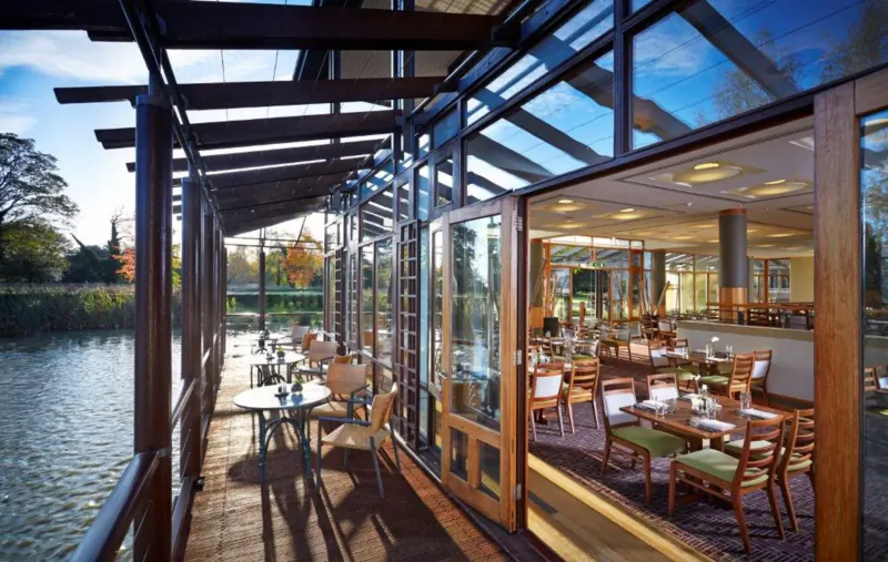 Glass-walled terrace restaurant at Fanhams Hall Hotel overlooking lake with tables, chairs, and autumn trees.
