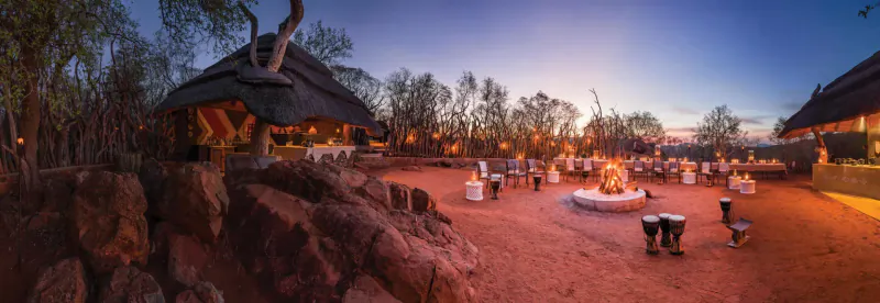 Panoramic sunset view of Madikwe Hills lodge: thatched buildings, fire pit, drums, guests on red sandy ground amid rocks and trees.