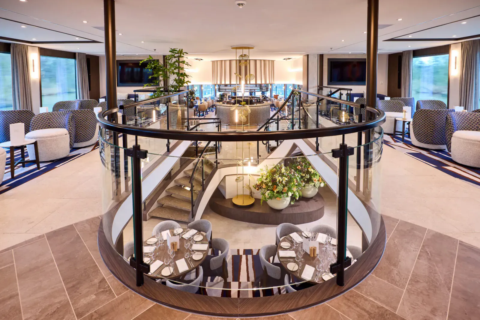 Elegant multi-level cruise ship atrium lounge with glass railings, round dining tables set for meals, plants, and large windows.