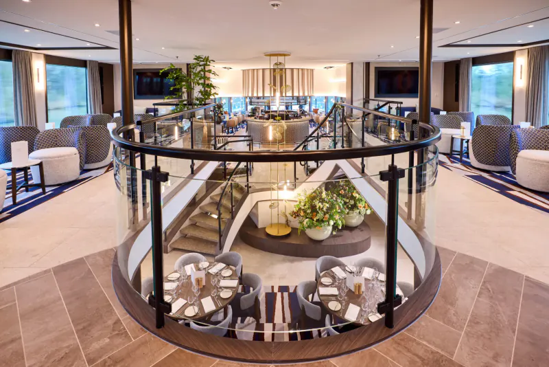Elegant multi-level cruise ship atrium lounge with glass railings, round dining tables set for meals, plants, and large windows.