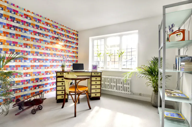 Cozy office in luxury cottage with colorful geometric wallpaper, yellow desk, chair, plants, bookshelves, and vintage toys.