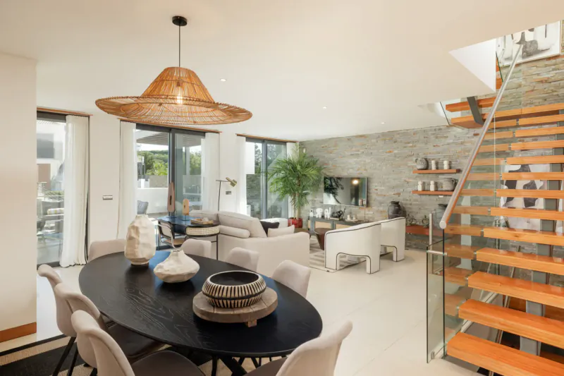 Spacious modern living-dining room in Vale do Lobo resort with rattan pendant light, oval table, ceramics, sofa, plants, and glass staircase
