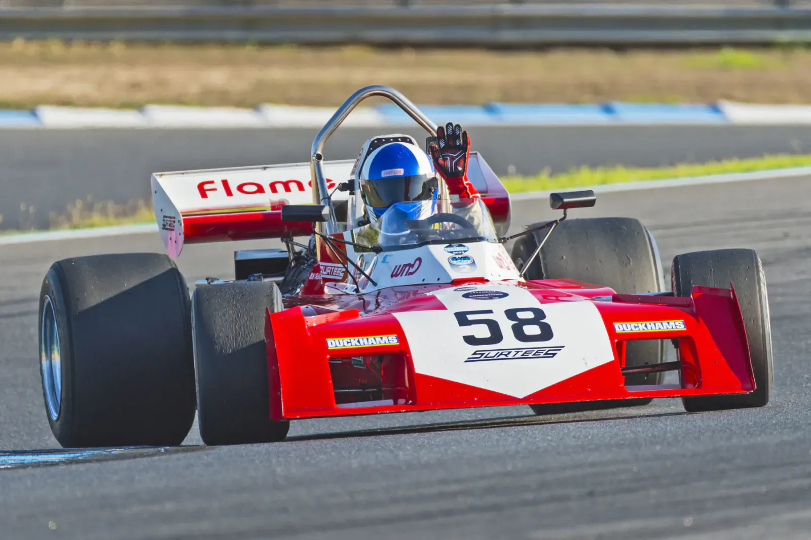 Red 'Flame' #58 racing car speeding on track, blue-helmeted driver waves gloved hand.