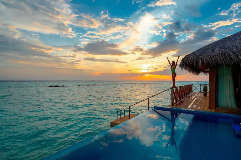 Woman in green bikini raises arms joyfully on overwater villa deck at tropical sunset with turquoise sea