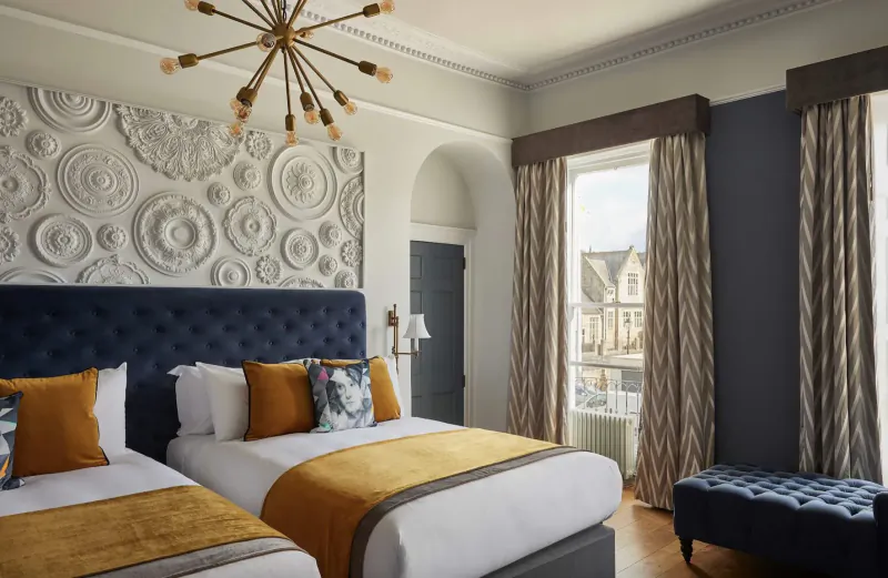 Elegant hotel room with navy upholstered twin beds, mustard pillows and throws, chandelier, ornate wall, and window view.