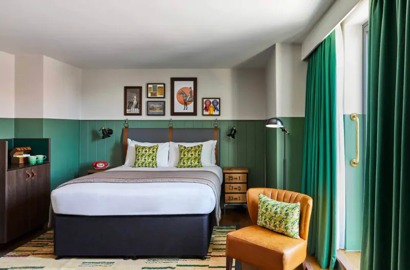 Cozy Hotel Indigo Chester room with green-accented walls, king bed with pillows, art frames, lamp, side tables, leather chair, and emerald curtains.