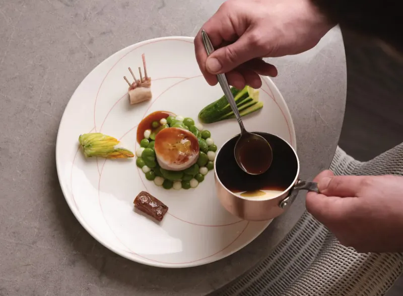 Chef pouring sauce from copper pot over plated dish with custard, peas, herbs, and haggis in cooking masterclass
