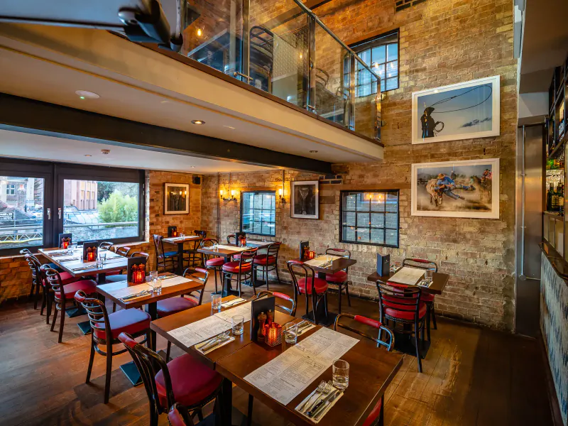 Cozy upscale restaurant interior with brick walls, red chairs, wooden tables set for dining, and canal view.