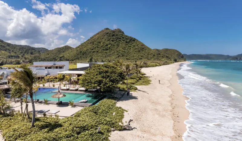 Aerial view of Amber Lombok Beach Resort with infinity pool, white sand beach, turquoise ocean, and lush green hills under blue sky.