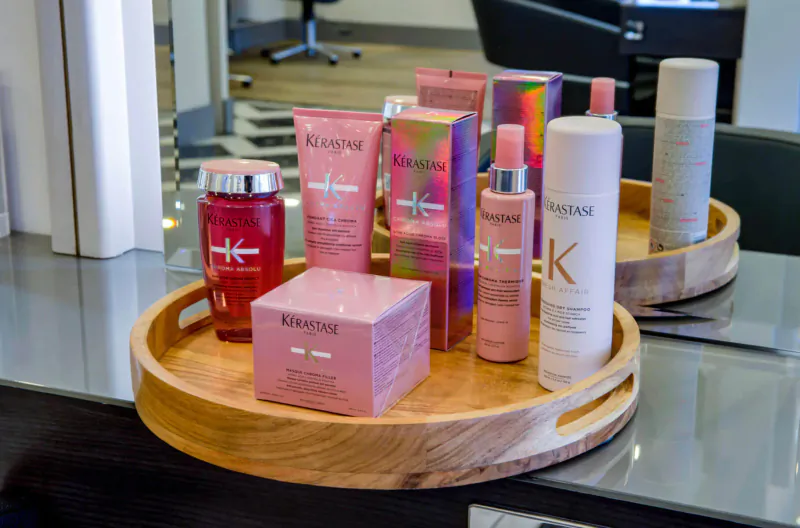 Kérastase hair products on wooden tray in salon: pink bottles, jars, boxes on glass counter with mirror reflection.